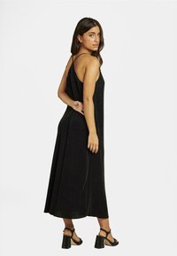 Robe noire sans manches avec une coupe ample, en tissu texturé et d'une longueur jusqu'aux chevilles, associée à des sandales à talons noirs avec un talon carré.