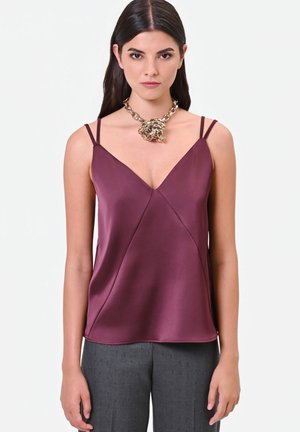 Femme aux cheveux raides et foncés porte un haut en satin bordeaux sans manches et un collier chaîne épais en or avec un détail nœud.