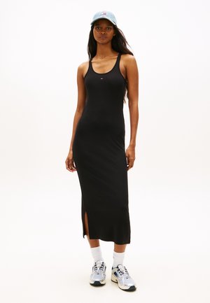Kobieta ubrana w czarną, bezrękawową, midi sukienkę z bocznym rozcięciem, jasnoniebieską czapkę, białe skarpetki i srebrne sneakersy, stojąca na białym tle.
