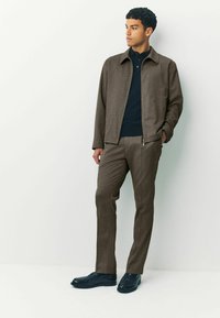 Giacca marrone con texture sopra un polo scuro, abbinata a pantaloni marroni coordinati e scarpe nere. Linee pulite, vestibilità moderna ed elementi di design sottili.
