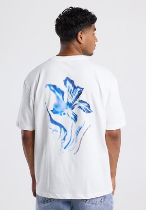 Witte katoenen t-shirt met een blauw bloemenpatroon op de achterkant en de tekst "De Botanische Tuin, Central Park, NY." Ontspannen pasvorm.