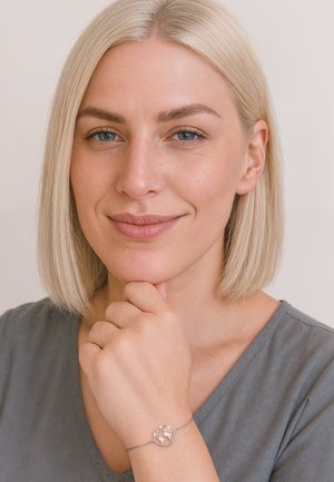 Srebrna bransoletka z okrągłym zawieszeniem przedstawiającym glob, noszona na ręce. Osoba nosząca ma krótkie blond włosy i jest ubrana w szary top.
