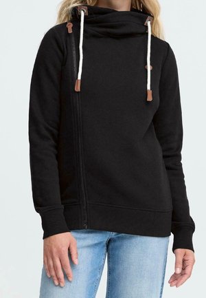 Femme portant un sweat à capuche noir avec des cordons blancs et une fermeture éclair asymétrique, associée à un jean bleu clair.