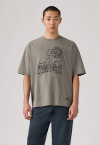 Νεαρός άνδρας που φοράει γκρι φαρδιά μπλούζα με μαύρο τριαντάφυλλο και τη λέξη «Permanent», συνδυασμένη με σκούρο μπλε τζιν, στέκεται μπροστά σε απλό φόντο.