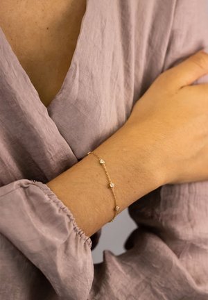 Goldene Kettenarmband mit drei kleinen klaren Edelsteinen, getragen an einer Hand auf einem hellen, strukturierten Mauve-Stoff.