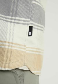 Gros plan de l'ourlet d'une chemise à carreaux gris, beige et blanc sur un pantalon gris, montrant la texture du tissu et une petite étiquette noire sur le côté de la chemise.