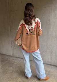 Hosszú hajú nő, aki világosbarna és fehér pulóvert visel, amelyen cowboy és ló mintája van, világoskék bő szárú farmert és világosbarna magassarkút, beltéren áll.