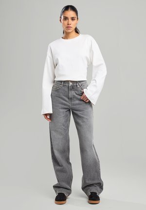 Witte lange mouwen top met een ronde halslijn, gecombineerd met hooggesnoerde, lichtgrijze wijde jeans. Zwarte sneakers met bruine zolen.