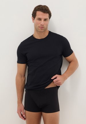 Man draagt een nauwsluitend zwart T-shirt en zwarte boxershorts, tilt zijn shirt iets op terwijl hij staat tegen een witte achtergrond.