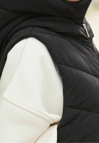 Czarna ocieplana kamizelka na białym swetrze o gładkiej fakturze. Posiada wysoki kołnierz i zamek błyskawiczny oraz pikowany wzór na kamizelce.