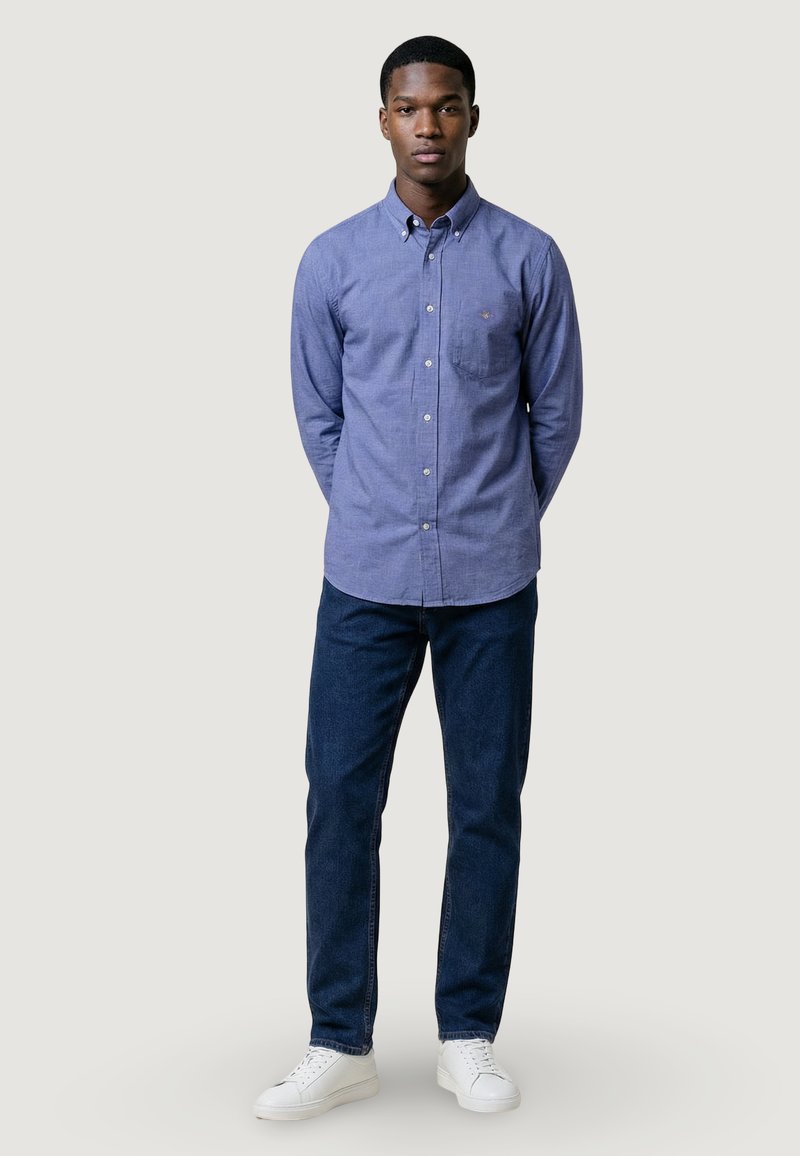 Jeune homme debout, les mains derrière le dos, portant une chemise bleue boutonnée, un jean foncé et des baskets blanches, sur un fond uni.