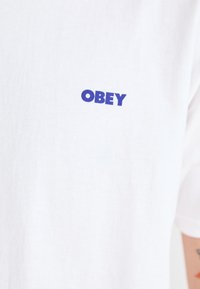White cotton t-shirt featuring a blue "OBEY" logo. The material is smooth and lightweight, with a relaxed fit and short sleeves.