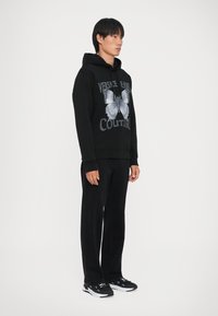 Hanorac negru cu imprimeu de fluture și text "VERSACE ENSEIGN", asortat cu pantaloni negri drepti și adidași negri.