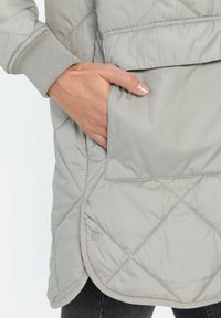 Grau gesteppte Jacke mit abgerundetem Saum, gerippten Bündchen und einer sichtbaren Seitentasche. Der Stoff wirkt glatt und leicht glänzend.