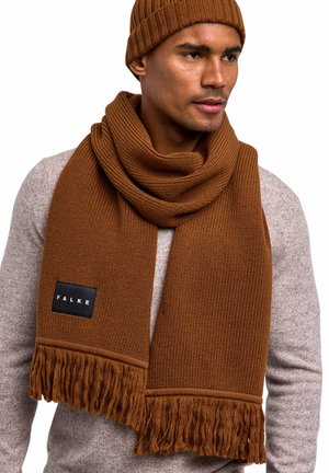 Sciarpa in maglia marrone con dettagli frangiati e un'etichetta nera, accompagnata da un berretto a coste coordinato, indossata sopra un maglione grigio chiaro.