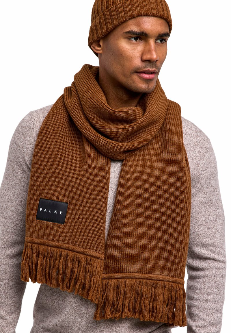 Écharpe en tricot marron avec des franges et une étiquette noire, accompagnée d'un bonnet à côtes assorti, portée sur un pull gris clair.