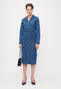 Μπλε πουκαμίσα-φόρεμα από τζιν με κουμπιά μπροστά, μακριά μανίκια, ζώνη στη μέση και κανονικό γιακά. Συνδυάστε με μαύρα παπούτσια και τσάντα.