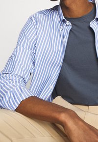 Blauw-wit gestreept overhemd met lange mouwen over een donkergrijs T-shirt. Lichtbeige broek. Katoenen stof met knoopdetails en opgerolde mouwen.