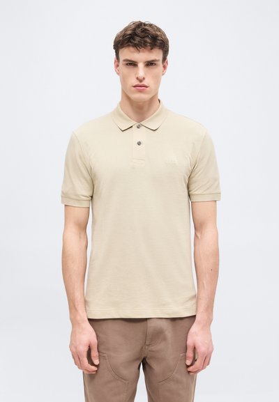 Jeune homme de face portant un polo beige à manches courtes et un pantalon marron, sur un fond clair uni.