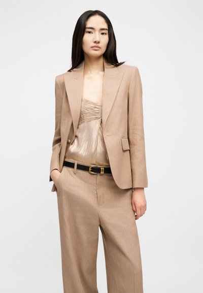 Γυναίκα με μπεζ κοστούμι με λεπτές ρίγες, που αποτελείται από ταιριαστό παντελόνι και σακάκι, από πάνω φοράει σατέν τοπ με ρυτιδωτή υφή, τα χέρια της είναι μερικώς στις τσέπες, με ουδέτερη έκφραση.