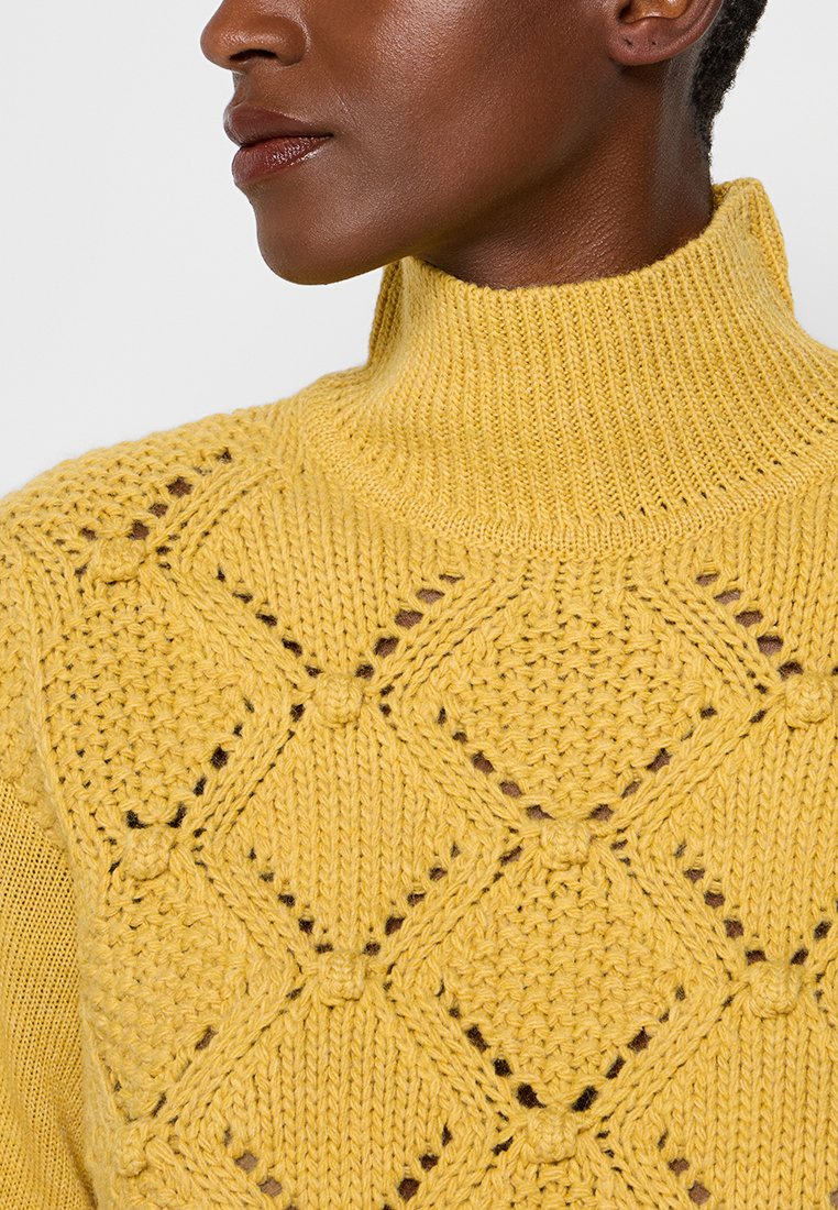 Maglione giallo a collo alto lavorato a maglia, caratterizzato da un intricato motivo a diamante con dettagli a pompon e un collo a coste. Texture morbida.