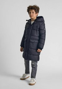 Zateplená čierna parka s kapucňou, predným zipsom, dvoma vreckami a rebrovanými manžetami; nosená cez biele tričko a šedé roztrhnuté džínsy.