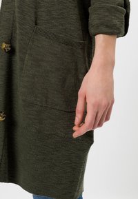 Dlouhý olivově zelený kardigan z texturované pletené látky, který má zapínání na knoflíky a přední kapsu na levé straně.