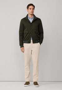 Uomo in giacca bomber verde oliva, camicia a righe blu, pantaloni beige e sneakers marroni, in piedi di fronte a uno sfondo grigio chiaro uniforme.