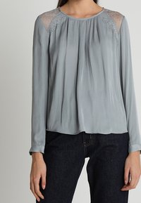 Das Model trägt eine hellgraue Bluse mit Spitzenakzenten an den Schultern, kombiniert mit dunklen Jeans. Sie steht vor einem neutralen Hintergrund.