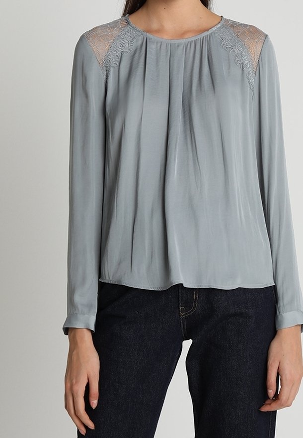 Das Model trägt eine hellgraue Bluse mit Spitzenakzenten an den Schultern, kombiniert mit dunklen Jeans. Sie steht vor einem neutralen Hintergrund.