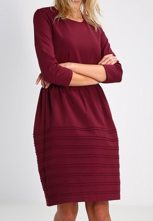 Vrouw in een knielange, bordeauxrode jurk met lange mouwen en horizontale plooien op de rok, staand met gekruiste armen.