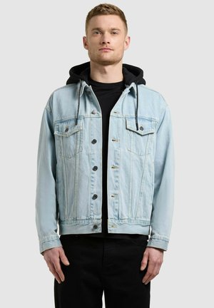 Mladý muž v svetlomodrej rifľovej bunde cez čiernu mikinu s kapucňou a čierne nohavice, stojaci pred jednoliatym svetlosivým pozadím.