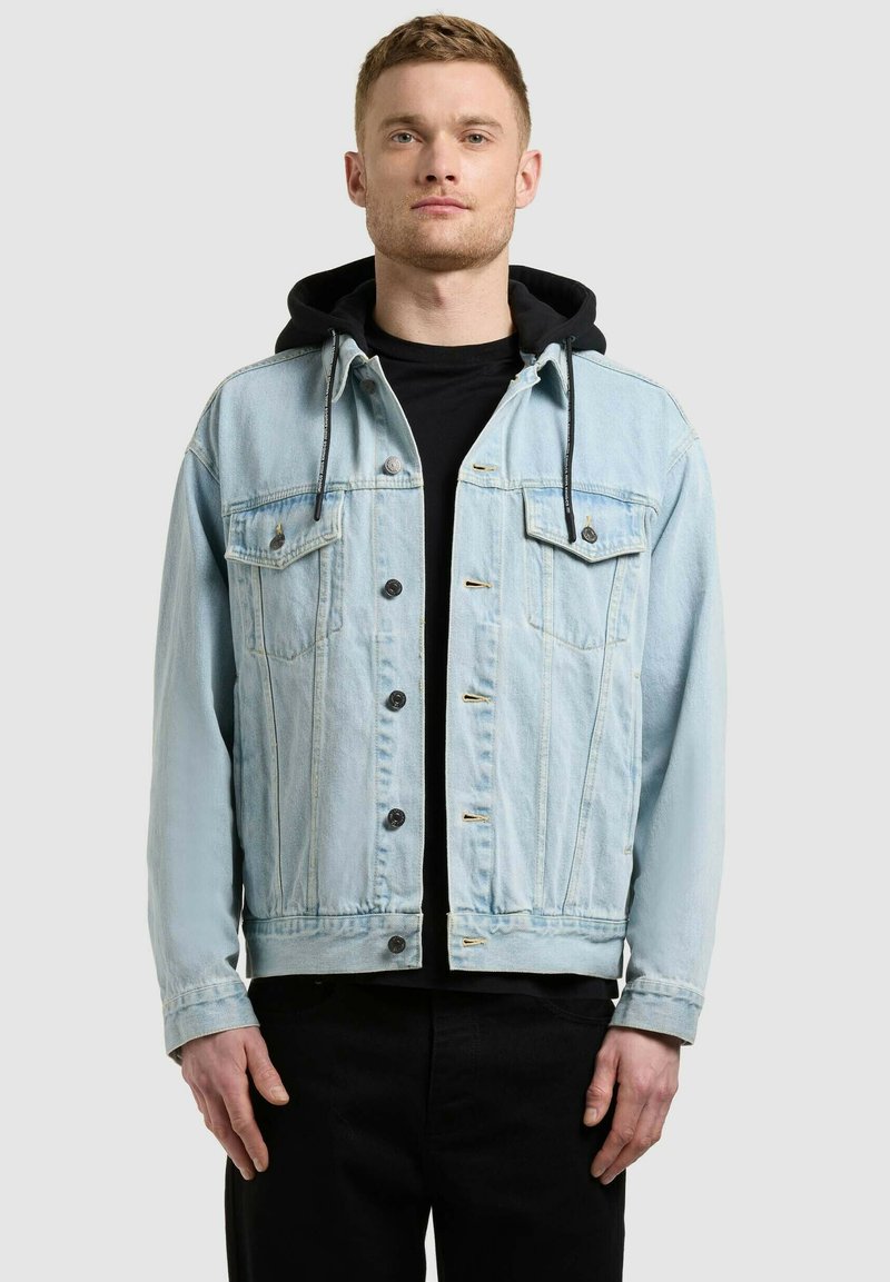 Jeune homme portant une veste en denim bleu clair par-dessus un sweat à capuche noir et un pantalon noir, debout devant un fond gris clair uni.