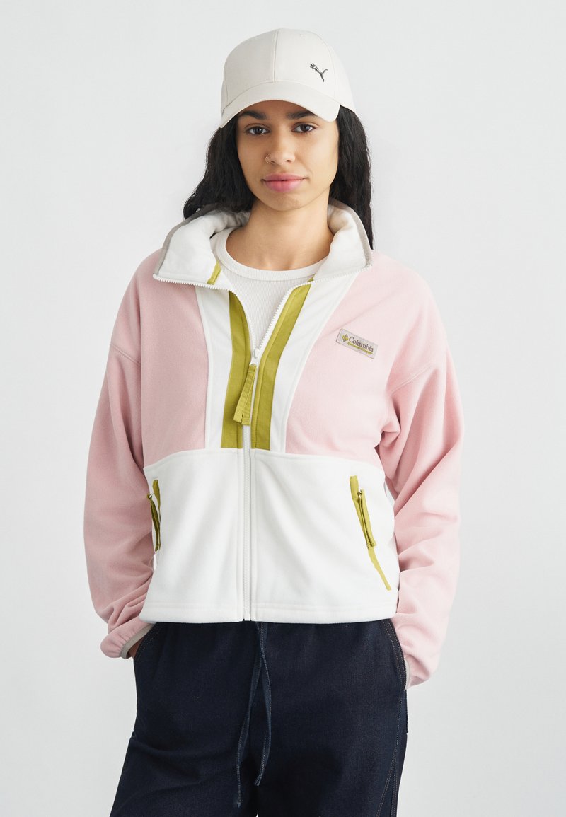 Φούξια ροζ και λευκό fleece μπουφάν με ψηλό γιακά, πράσινες λεπτομέρειες φερμουάρ και δύο τσέπες με φερμουάρ. Φορεμένο πάνω από λευκή μπλούζα.