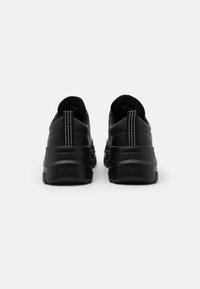 Zapatillas negras con una parte superior de cuero liso, costuras blancas prominentes y una suela de goma gruesa. Cuenta con una lengüeta en la parte posterior para facilitar el uso.