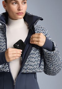 Pletena plava i bijela skraćena jakna s visokim ovratnikom i unutarnjim patentnim zatvaračem, uparena s kremastim topom s lažnim ovratnikom.