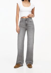Jean gris à jambes larges avec taille haute, effet délavé et design classique à cinq poches. Associé à un crop top blanc ajusté et des chaussures marron.