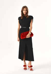 Robe noire sans manches avec une fente frontale, taille ceinturée, pochette en velours rouge avec détail floral, et sandales à talons rouges.