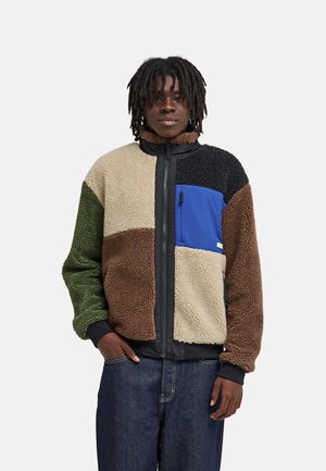 Jong persoon met dreadlocks draagt een kleurblok fleecejas met groene, bruine, beige, zwarte en blauwe panelen, staand met handen in de zakken.
