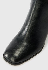 Juodi kulkšnies batai, pagaminti iš tekstūrinės sintetinės odos, su kvadratine nosimi ir glotnia apdaila. Padas juodas su subtiliais akcentais.