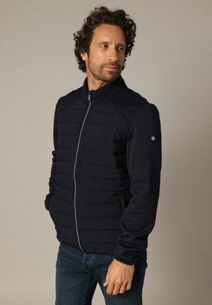 Veste marine avec fermeture éclair à l'avant, panneaux matelassés et textures côtelées aux épaules et aux manches. Elle est dotée de poches latérales et d'un logo sur la manche.