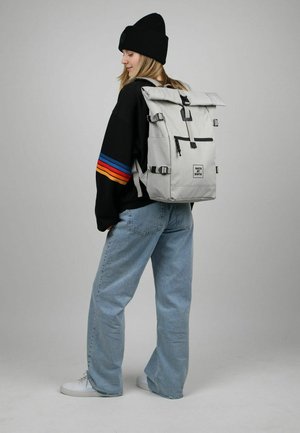 Light gray roll-top backpack with black hardware, adjustable straps, front pocket, and logo detail, paired with a black sweater and blue jeans.