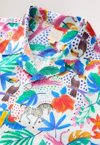 Kurzärmeliges Hemd aus leichtem Stoff mit buntem tropischem Muster mit Vögeln, Blumen und Blattwerk. Weiße Knöpfe sichtbar.