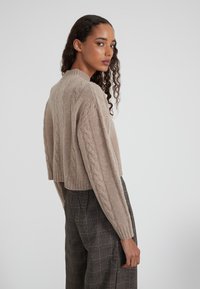 Hellbrauner, kurz geschnittener Kabelstrickpullover mit gerippten Akzenten und langen Ärmeln, kombiniert mit karierten Hosen. Strukturierte Garn-Details sind sichtbar.