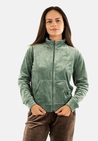 Giacca verde a zip realizzata in tessuto morbido e strutturato, con colletto alto e due tasche frontali, abbinata a pantaloni marroni.