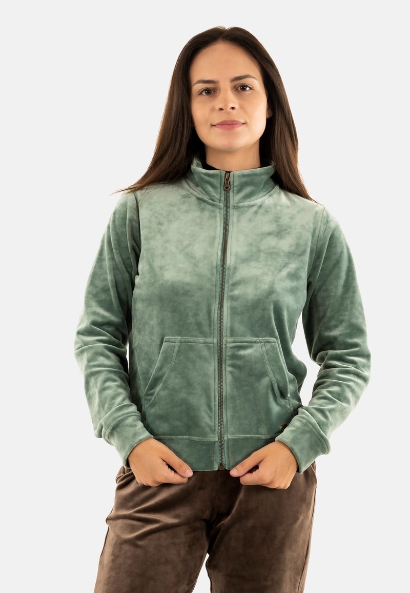 Giacca verde a zip realizzata in tessuto morbido e strutturato, con colletto alto e due tasche frontali, abbinata a pantaloni marroni.
