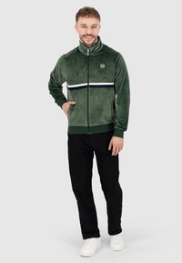 Giacca in velluto verde con zip e colletto alto, striscia bianca e blu sul petto e tasche con cerniera. Abbinata a pantaloni neri e sneakers bianche.