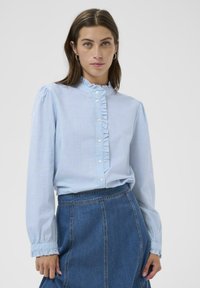 Chemise bleu clair à manches longues avec un col volanté et une patte de boutonnage à l'avant, dotée de boutons blancs et de poignets froncés, associée à une jupe en denim.