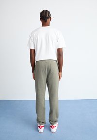 Bărbat cu părul împletit, îmbrăcat cu un tricou alb, pantaloni verde măsliniu și adidași albi cu accente roșii, stând pe un podea albastră, cu fața întoarsă.