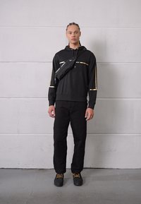 Sudadera negra con detalles dorados, combinada con pantalones negros y zapatos negros. Se lleva un bolso cruzado negro sobre el pecho.